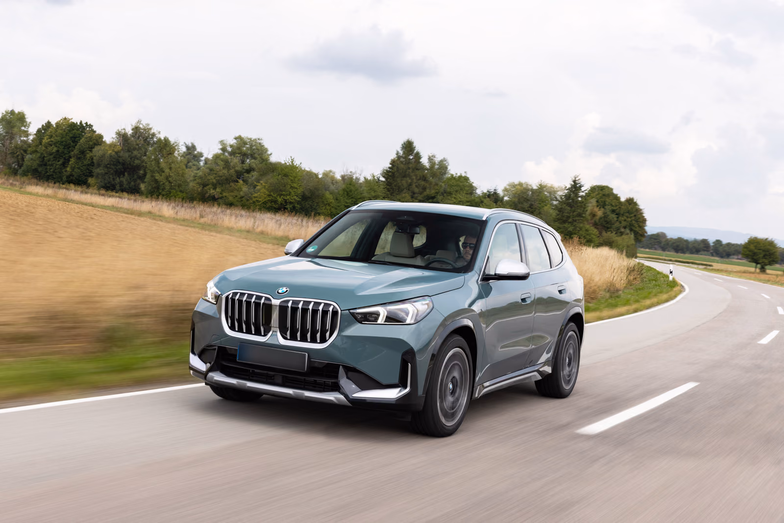 BMW X1 U11 — серо-зелёный кроссовер спереди в три четверти на загородной дороге